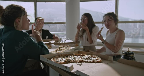 Multicultural business team enjoys pizza in a modern office with large windows and a scenic cityscape view.