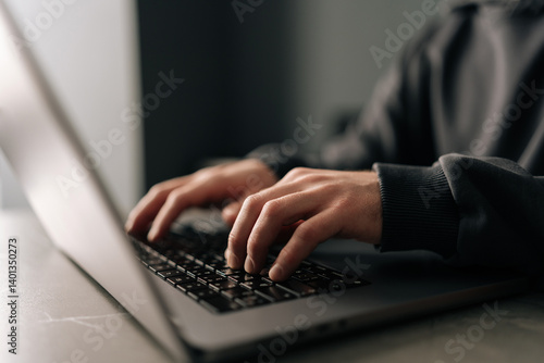 Close-up hands of unrecognizable programmer male typing on laptop keyboard, immersed in software development or coding, sitting at table by window in dark room. Concept of remote job, freelance.