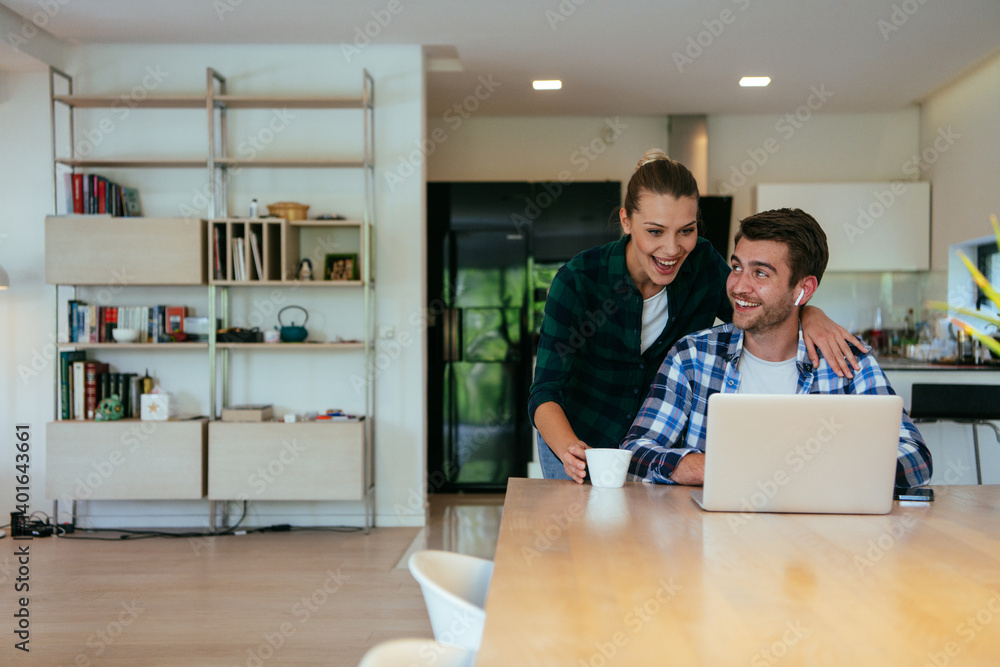 A young married couple is talking to parents, family and friends on a video call via a laptop while sitting in the living room of their modern house.