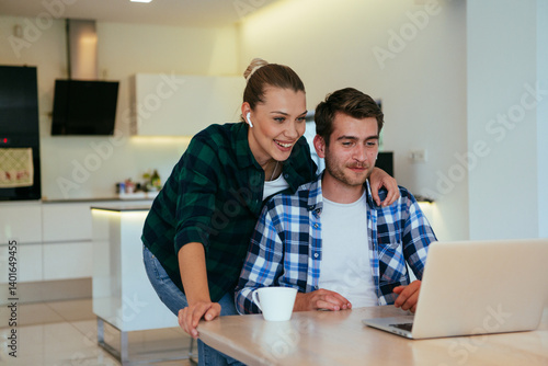 A young married couple is talking to parents, family and friends on a video call via a laptop while sitting in the living room of their modern house.