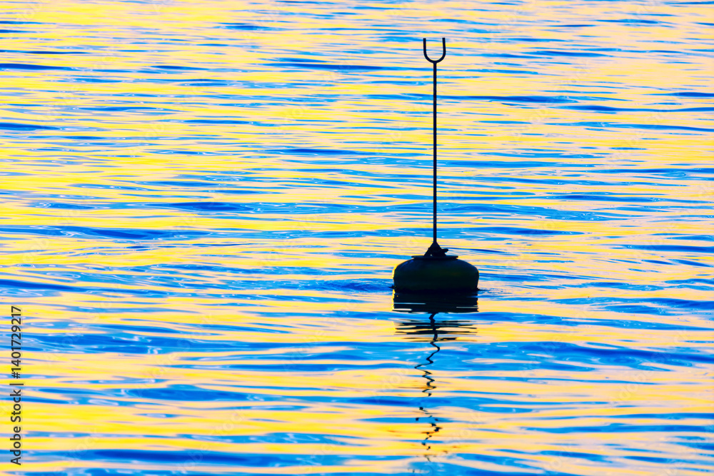 Lone buoy with a tall marker floats on a shimmering ocean, reflecting hues of blue and gold from the setting sun. Serene water surface creates a minimalist scene