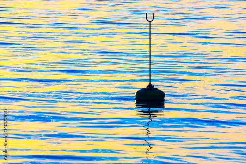 Lone buoy with a tall marker floats on a shimmering ocean, reflecting hues of blue and gold from the setting sun. Serene water surface creates a minimalist scene