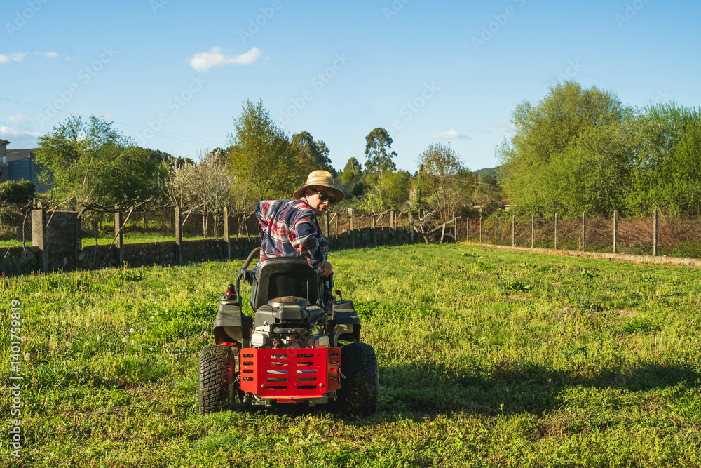 Obraz premium Man mowing spring lawn on red tractor in garden with apple and pear trees. Wearing work boots and flannel in rural setting.