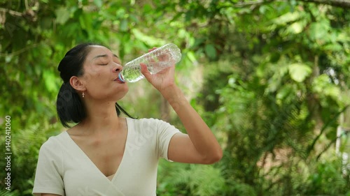 Woman staying hydrated by drinking from a plastic bottle in a lush green forest. Refreshing break during an outdoor trip.
