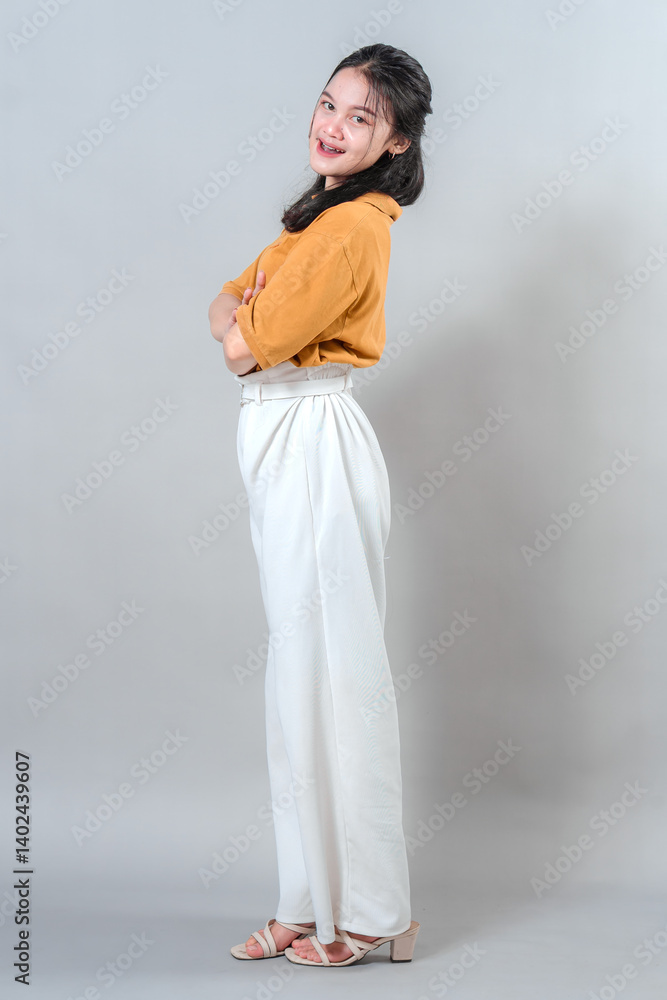 Confident young woman smiling sweetly with gentle facial expression, arms crossed and body slightly turned in relaxed pose, portraying calmness and friendliness in studio shoot.