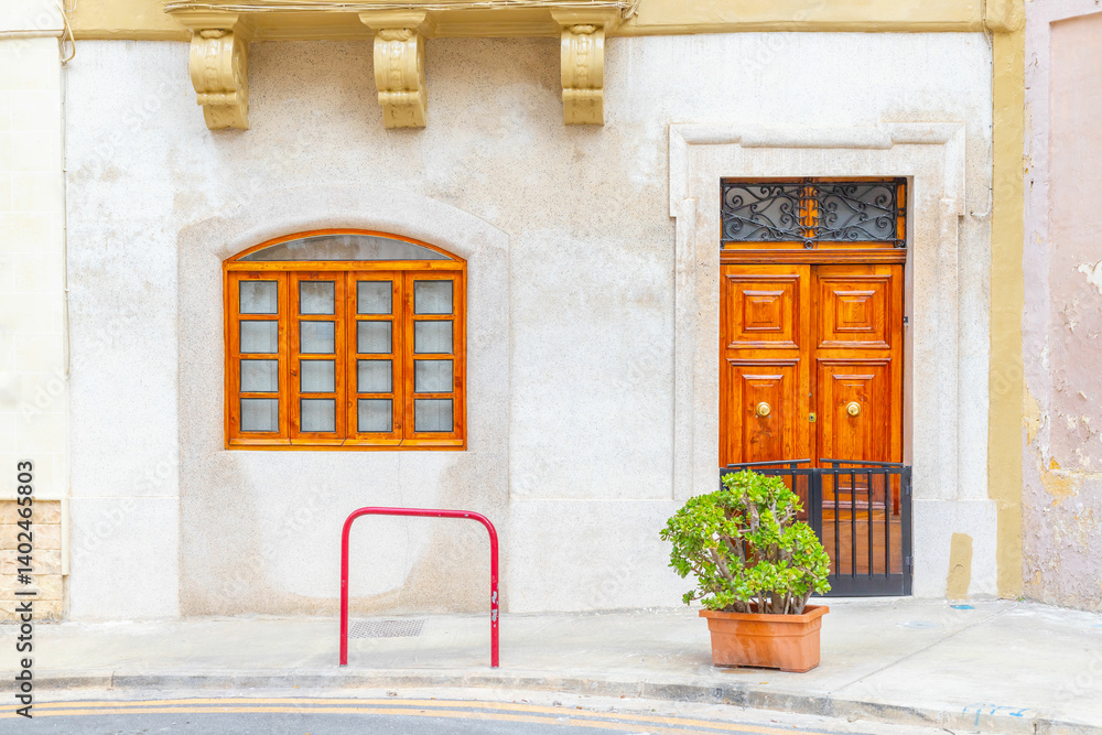 Facade of an old Maltese residential house with a brown wooden door, a window and a potted plant in front of it