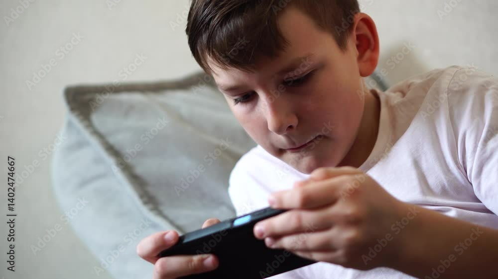 A ten-year-old boy in a white T-shirt enthusiastically plays on his phone while lying on his bed. He leaned on one elbow A ten-year-old boy in a white T-shirt enthusiastically plays on his phone while lying on his bed. He leaned on one elbow