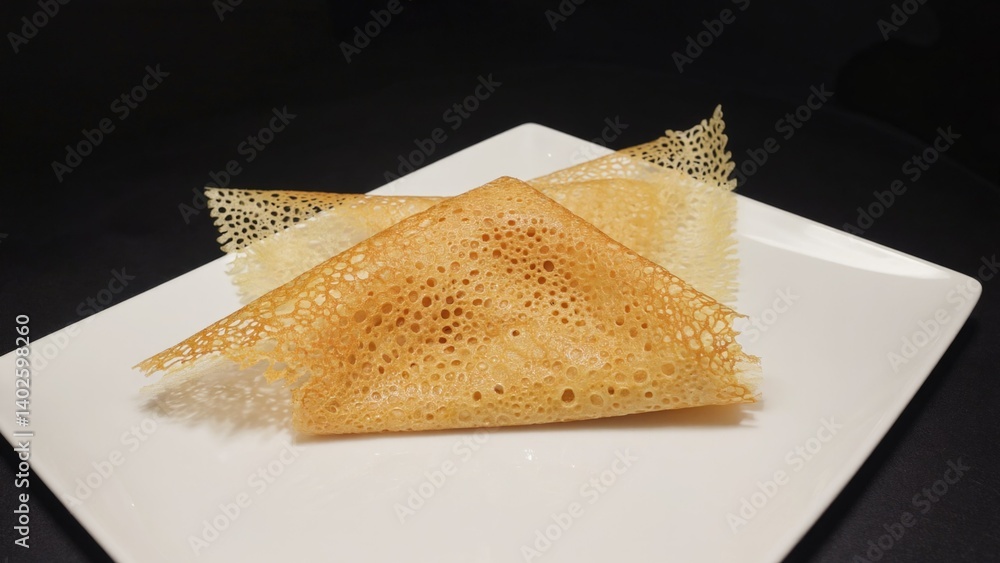 An artfully stacked crepe with intricate holes, presented on a white square plate for a gourmet experience.