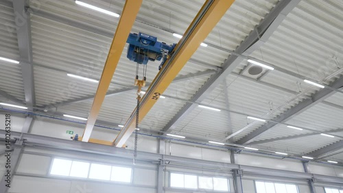 An industrial yellow overhead crane with a blue hoist system is suspended from steel beams, ready for heavy lifting operations in a clean, modern manufacturing facility