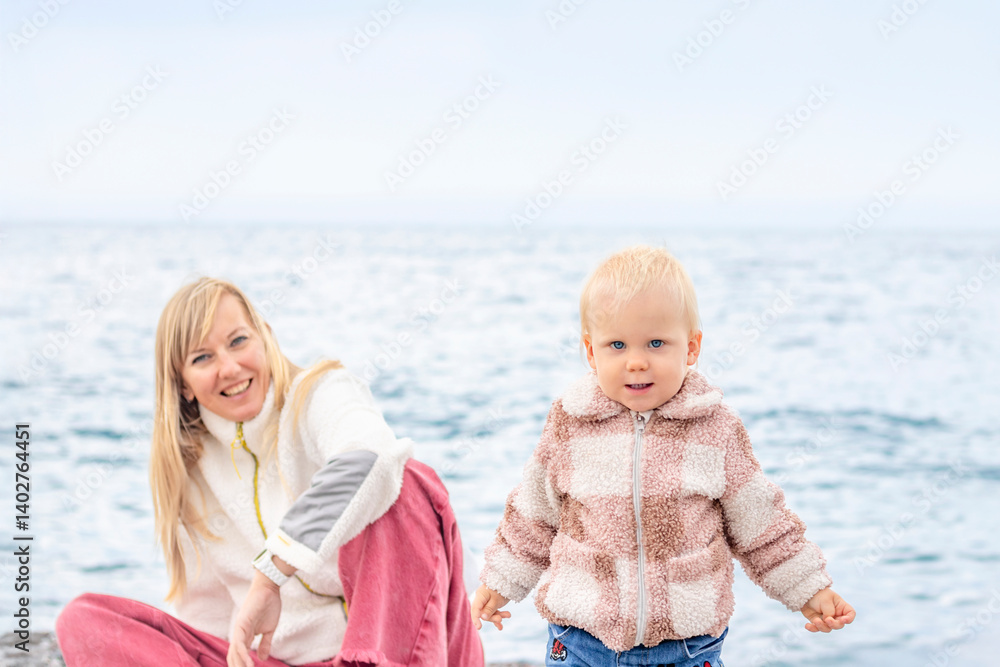 A joyful mother and her child play on the beach, surrounded by pebbles with the sea in the background.