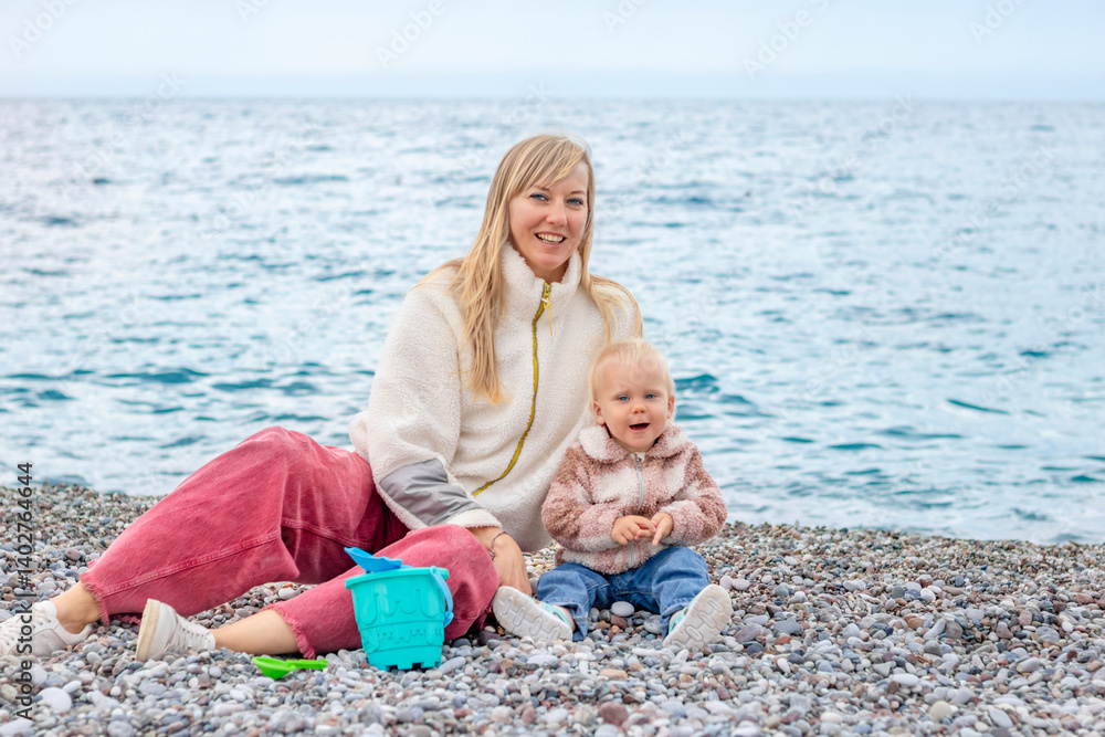 A joyful mother and her child play on the beach, surrounded by pebbles with the sea in the background.