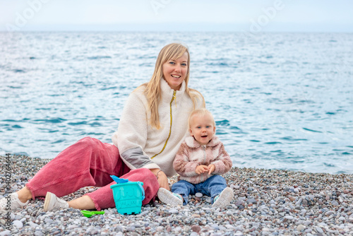 Obraz na płótnie A joyful mother and her child play on the beach, surrounded by pebbles with the sea in the background. w salonie