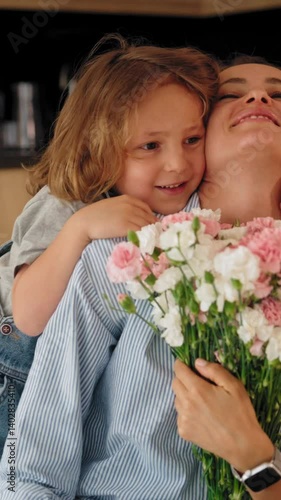 Young boy gives his mother a bouquet of flowers at home in a warm and cozy setting. She smiles with joy and gratitude. Concept of Mother's Day celebration and family love. Mid-range shot natural light