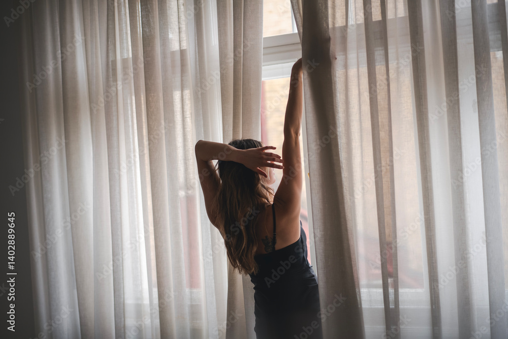 Young slim woman in black dress opening white curtains standing at window
