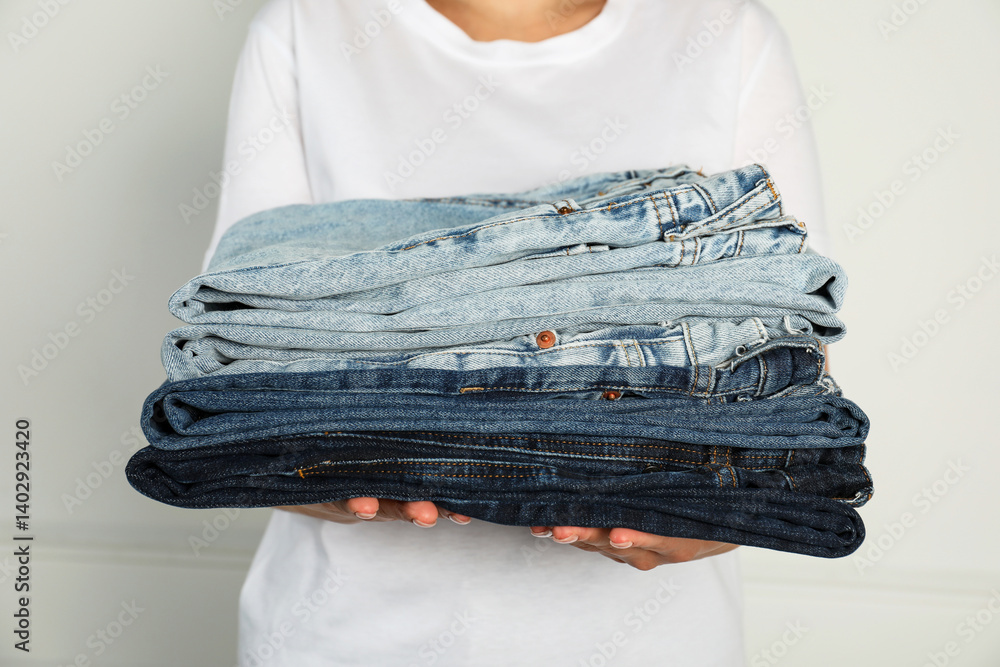 Woman with stack of different stylish jeans near light wall indoors, closeup