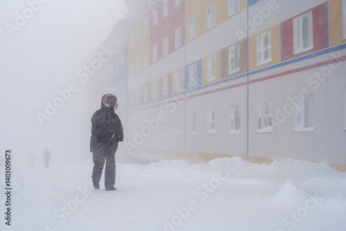 Blizzard in the city. A man walks along a snowy street past a residential building. Snowstorm with strong wind. Harsh polar climate in the Arctic. Cold winter weather. Anadyr city, Chukotka, Russia.