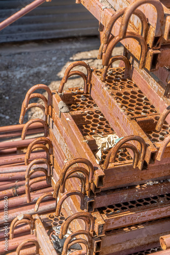 This image displays a tightly stacked arrangement of rusty steel scaffolding components at a construction site. The perforated steps and U-shaped hooks are clearly visible, showing signs of wear 