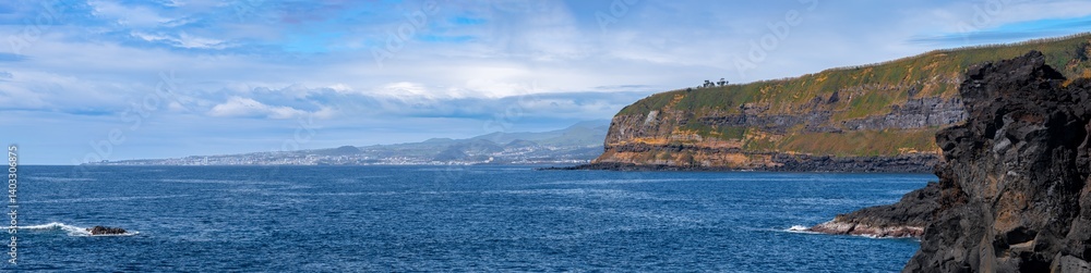 Fototapeta premium Blick von Água de Pau in Richtung Ponta Delgada (Azoren). Im Vordergrund massive Lavafelsen und blaues Meer, dazwischen die Bucht Baixa d'Areia Beach. Im Hintergrund Ponta Delgada unter blauem Himmel.
