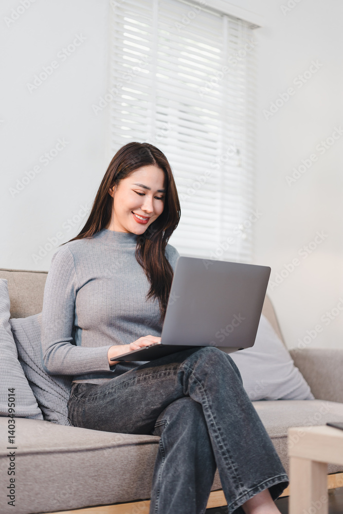 Asian businesswoman sitting on sofa, smiling while working on laptop in bright and cozy living room Asian businesswoman sitting on sofa, smiling while working on laptop in bright and cozy living room