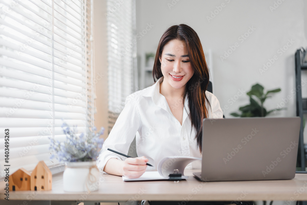Asian businesswoman smiling while working on laptop and taking notes in bright modern office Asian businesswoman smiling while working on laptop and taking notes in bright modern office