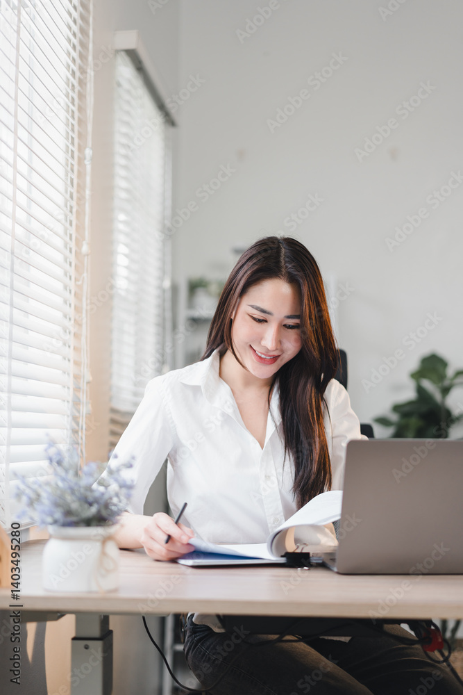 cheerful Asian businesswoman working at her desk with laptop, writing notes, and reading documents in bright office cheerful Asian businesswoman working at her desk with laptop, writing notes, and reading documents in bright office