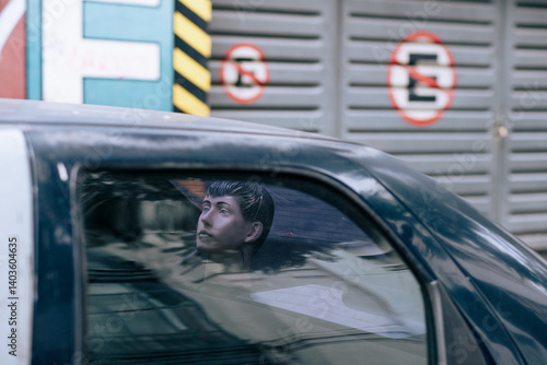 A plastic head man is gazing out of the window of a police van as it slowly drives through the city streets, observing the surroundings and people