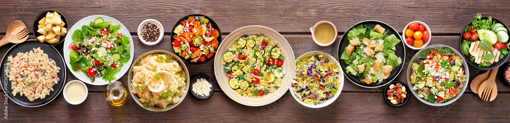 Assortment of delicious homemade salads. Overhead view table scene on a dark wood banner background. Summer food concept. Potato, caesar, pasta and green salads.