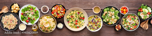 Obraz na płótnie Assortment of delicious homemade salads. Overhead view table scene on a dark wood banner background. Summer food concept. Potato, caesar, pasta and green salads. w salonie