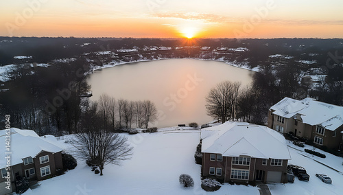 Snowy Suburban Neighborhood Overlooks Winter Lake at Golden Sunset with Dark Trees Silhouettes and Residential Buildings during Day in Wide Aerial Shot