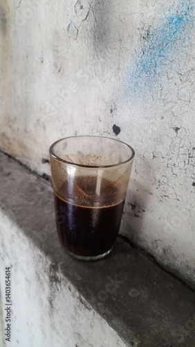 A small glass containing dark black coffee, placed on a narrow ledge against a rough, weathered wall with peeling paint. The coffee's dark color contrasts with the wall's texture