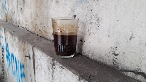 A small glass containing dark black coffee, placed on a narrow ledge against a rough, weathered wall with peeling paint. The coffee's dark color contrasts with the wall's texture