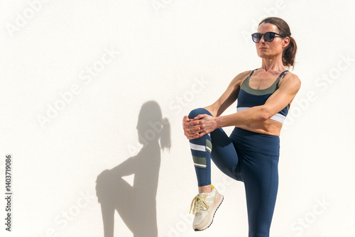 Sporty woman doing a leg stretch warm up exercise against a white wall.