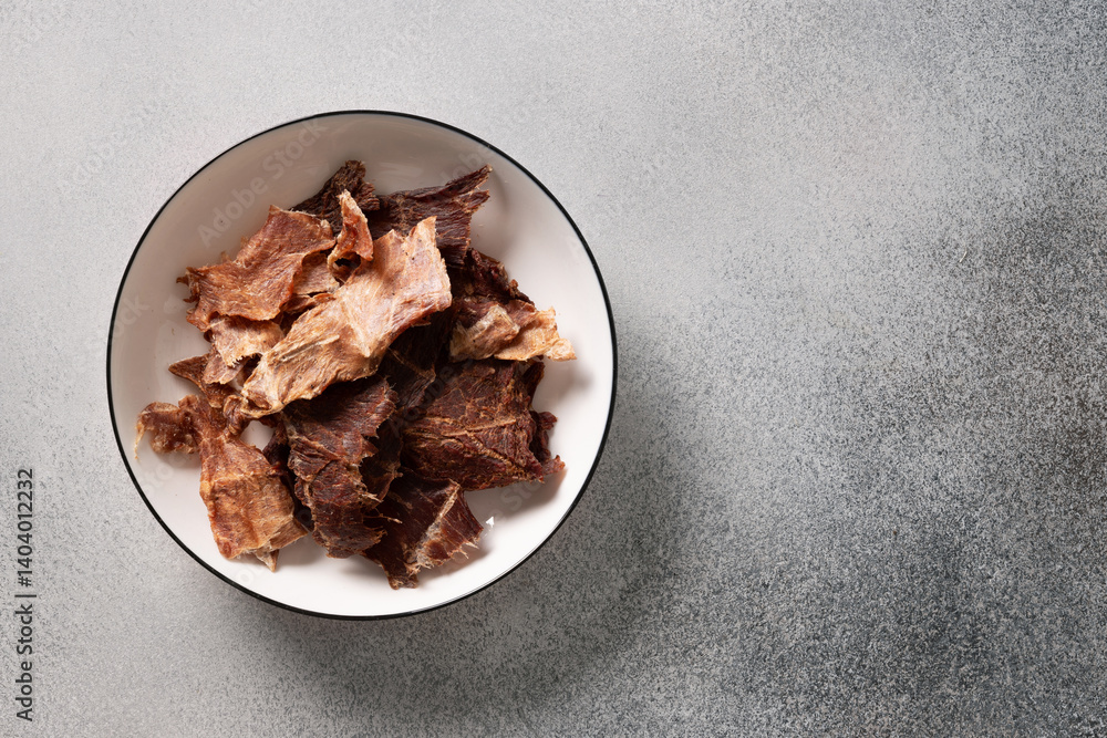 Bowl filled with savory beef jerky on a textured gray surface. Perfect for high-protein snack ideas, exploring flavors, hiking, camping, or healthy eating themes.