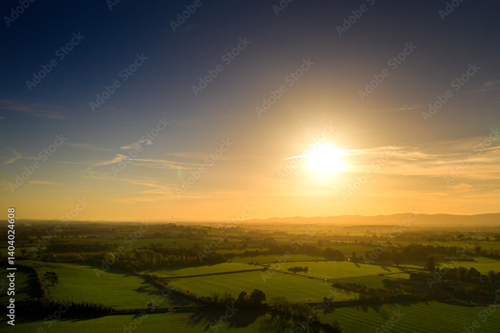 Fototapeta premium View of lush green farmland at golden hour, with long shadows cast by hedgerows and trees, glowing fields, and distant hills softly lit by the warm setting sun under a clear sky.