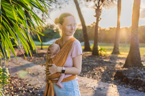 A loving mother walks through a peaceful park, carrying her baby securely in a sling. A heartwarming moment of maternal bonding, babywearing, and the joy of motherhood in nature