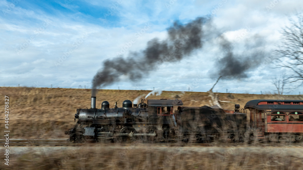 Obraz premium A vintage steam locomotive puffs black smoke as it moves along the tracks in a scenic countryside setting. The overcast sky adds a moody atmosphere to the scene.