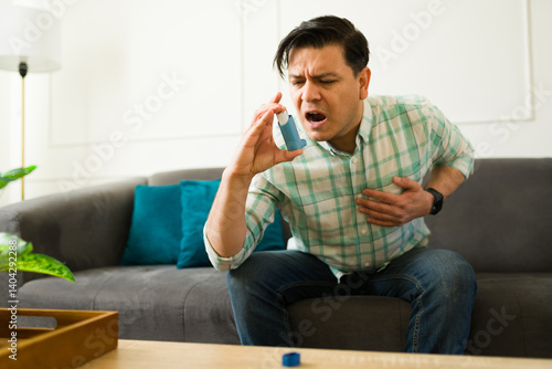 Man having breathing problems using an inhaler while sitting on sofa at home suffering asthma attack