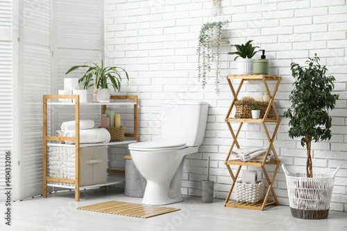 Interior of modern restroom with ceramic toilet bowl, houseplant and shelving unit near white brick wall