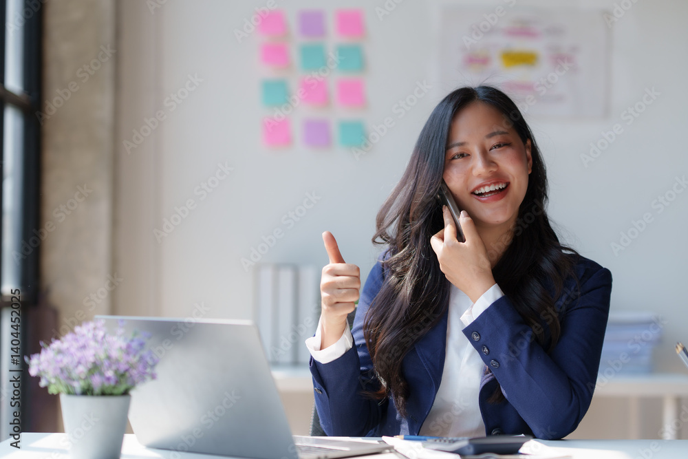 Cheerful businesswoman gesturing thumbs up while speaking on smartphone and working on laptop in modern office, expressing positivity and success Cheerful businesswoman gesturing thumbs up while speaking on smartphone and working on laptop in modern office, expressing positivity and success