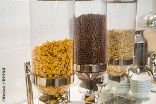 Close-up view of cereal dispensers filled with cornflakes, chocolate balls, and oats in a breakfast buffet setting