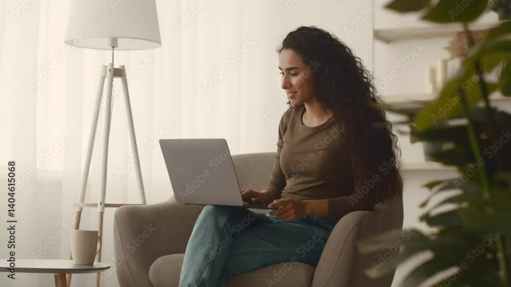 A young Middle Eastern woman sits comfortably in a cozy chair, focused on her laptop. Natural light fills the room, creating a relaxed atmosphere perfect for leisure and productivity. A young Middle Eastern woman sits comfortably in a cozy chair, focused on her laptop. Natural light fills the room, creating a relaxed atmosphere perfect for leisure and productivity.
