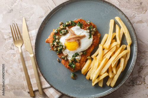 Delicious breaded schnitzel with anchovies, fried egg and capers parsley sauce on top served with french fries close-up on a plate on the table. Horizontal top view from above