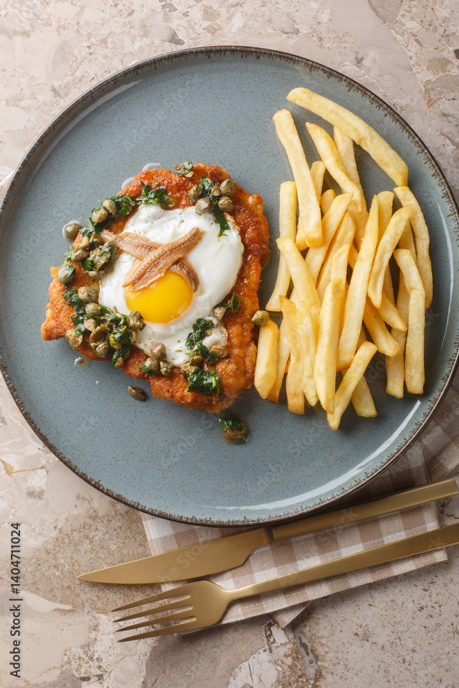 Schnitzel Holstein topped with a fried egg, anchovies, capers and lemon parsley brown butter served with fries close up on the plate on the table. Vertical top view from above
