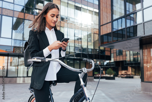 Young businesswoman sitting on a bicycle and checking her smartphone. Positive female entrepreneur with bicycle in courtyard of business building.