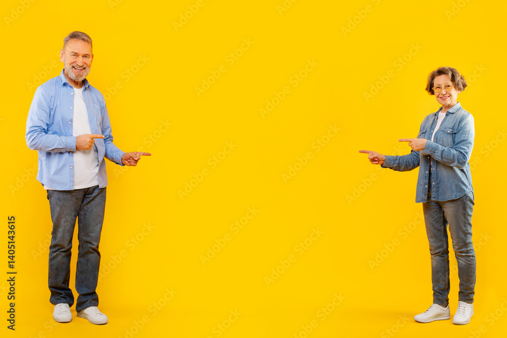 Cheerful senior couple pointing fingers at empty space between them, showing great sale offer or promoting deal, standing on yellow background, full length Cheerful senior couple pointing fingers at empty space between them, showing great sale offer or promoting deal, standing on yellow background, full length