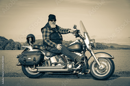 Kräftig gebauter Rocker sitzt auf einem klassischen, schweren Motorrad am Straßenrand, Sepia - Aufnahme.