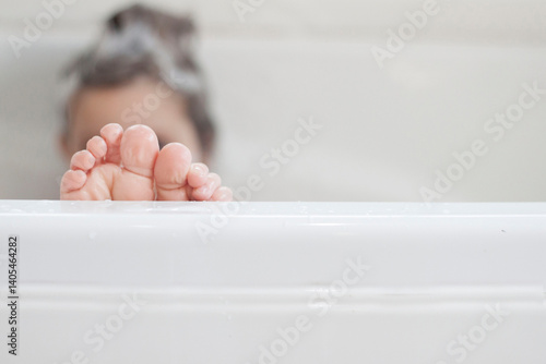 Girl sitting in a bath with shampoo in her hair and the focus on her bare feet sticking over the edge of the bathtub