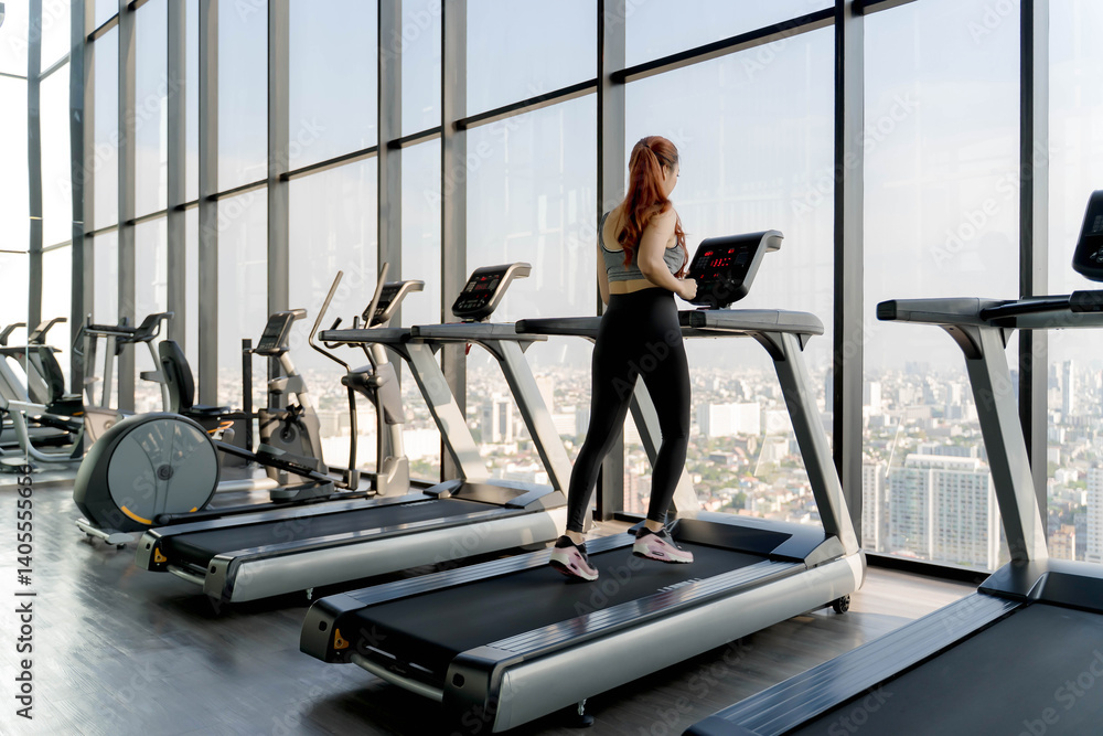Fototapeta premium Woman using treadmill in modern high-rise gym with panoramic city view, engaging in indoor cardio workout and healthy lifestyle fitness training routine