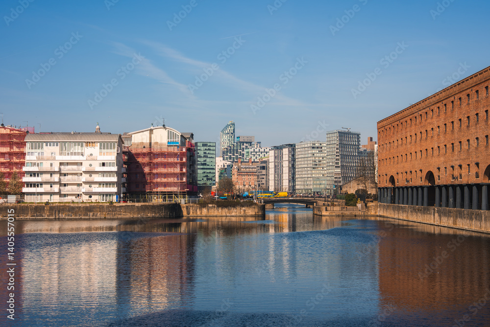 Fototapeta premium The image shows Liverpool's Albert Dock with red brick architecture, modern glass buildings in the background, calm water, and a small central bridge.