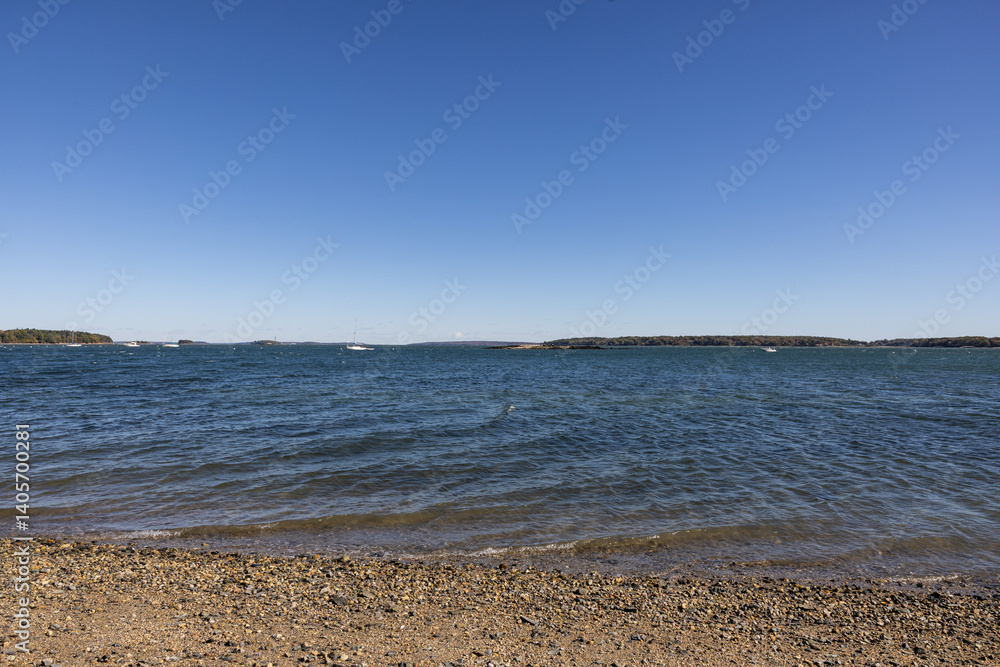 Photograph of pebble sandy shore with calm rippling waters on bright blue clear day Photograph of pebble sandy shore with calm rippling waters on bright blue clear day